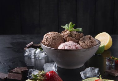 Bir kasede farklı renk ve dokulara sahip üç çeşit dondurma bulunur. Taze çilek, limon ve limon kâsenin yanına oturur. Çikolata ve buz parçaları karanlık yüzeye saçıldı..