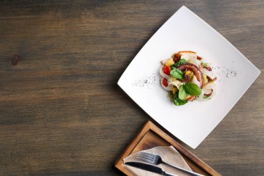 Ahşap bir masanın üzerinde duran kare beyaz bir tabakta renkli bir tabak sunulur. Tabak, ahşap bir tepside çatal bıçakla birlikte çeşitli malzemeler içeriyor..