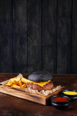 Siyah çörekli bir hamburger, ahşap bir tepside oturur. Hamburgerin üstünde erimiş peynir var. Onun yanında altın kızartmalar var. Tepsiye soslu üç küçük kase konur..