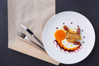 Yuvarlak beyaz bir tabak yuvarlak sarı bir yumurtanın yanında bir parça et tutar. Yemeğin etrafında soslar ve garnitürler var. Çatal bıçak takımı tabağın yanındaki masaya konur..