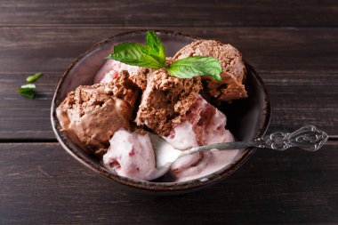 Bir kase dolusu çikolata ve çilekli dondurma içerir. Bir kaşık kasenin yanında duruyor. Dekor ahşap bir masa yüzeyine sahip. Dondurmanın üstüne taze nane yaprakları konur..
