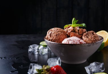 Dondurmanın çeşitli tatlarını içeren bir kase karanlık bir yüzeye oturur. Taze çilek ve nane yaprakları mevcut. Tabağın etrafına buz küpleri ve turunçgiller konacak..