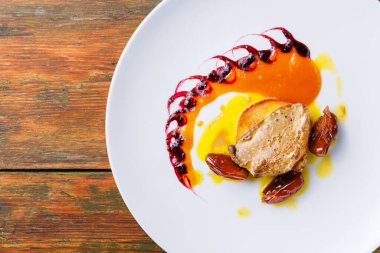 Bir şef, içinde renkli meyve sosu ve beyaz bir tabakta hurma bulunan bir yemek sunar. Dekor büyük ihtimalle ahşap masalı bir restoranda. Yemekler iyi düzenlenmiş görünüyor..