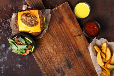 Bir dilim peynirli bir hamburger ahşap bir tahtanın üzerinde durur. Yanında salata ve patates dilimleri var. Yakınlarda küçük kaselerde soslar var..