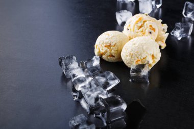 Karanlık bir yüzeyde üç kepçe dondurma dinleniyor. Kepçelerin etrafındaki buz küpleri temizdir. Sahne eğlenmeye hazır bir ziyafet gösteriyor. Dondurma, içinde küçük parçalar olan kremalı görünüyor..