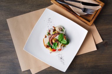 Bir deniz ürünleri tabağı, sebzeler de dahil olmak üzere çeşitli malzemelerle beyaz bir tabağa yerleştirilir. Yanında ahşap masa ve çatal bıçak bulunan bir restoranda servis ediliyor..