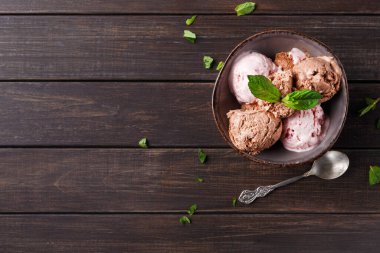 Bir kase üç farklı dondurma çeşidi içerir. Tahta masadaki kasenin etrafını taze nane yaprakları sarmış. Gümüş bir kaşık kasenin yanında duruyor..