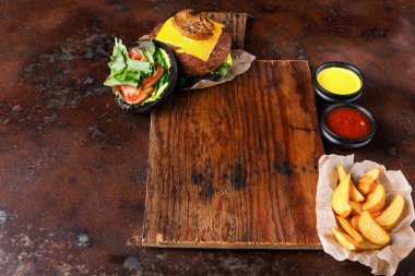 Bir hamburger, taze sebzeler ve iki küçük kase sosla birlikte ahşap bir tahtanın üzerinde oturur. Kızarmış patatesler hamburgerin yanına bir parça kağıt üzerine konur..
