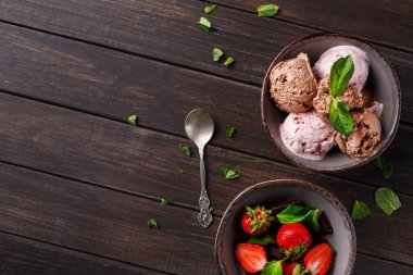 İki kase ahşap bir masanın üzerinde. Bir kasenin üzerinde farklı nane aromalı dondurma var. Diğer kasede taze çilek var. Kaselerin yanında gümüş bir kaşık var..