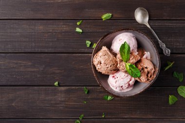 Bir kase dolusu çikolata ve çilekli dondurma içerir. Nane yaprakları en üste konur ve dağınık nane yaprakları ahşap masanın üzerindedir. Yakınlarda gümüş bir kaşık var.