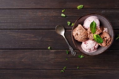 Bir kase üzerinde taze nane yaprakları olan üç çeşit dondurma tutar. Kase koyu ahşap bir masanın üzerine konur. Kasenin yanında bir kaşık yatıyor..