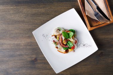 Salata beyaz bir tabağa konur. İçinde ızgara tavuk, sebze ve garnitür var. Çatal bıçak takımı tabağın yanında, tahta bir masanın üzerinde. Yemek ya da öğle yemeği için hazır görünüyor..