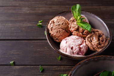 Bir kasede dört kepçe dondurma ahşap bir masanın üzerine konur. Nane yaprakları kasenin etrafına saçılmış. Çikolata, çilek ve vanilya aromalı..