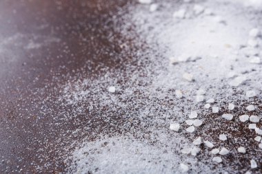 Beyaz tuz, siyah bir ahşap masaya eşit olmayan bir şekilde dağılmış. Tuz taneleri ışıkta hafifçe parıldıyor. Bu sahne mutfak aktivitesinin bir kısmını gösteriyor. Yemek pişirmek ya da baharat katmak..