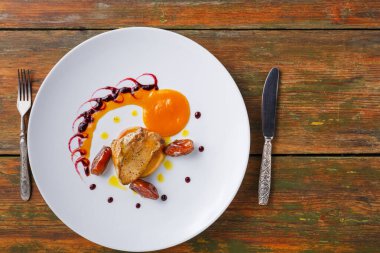 Bir gurme tabağı renkli soslar ve süslemelerle çevrili bir parça et içerir. Tabak, kenarlarında çatal bıçak takımı olan ahşap bir masaya konur..