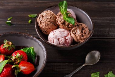 Çikolata ve çilekli dondurma kadehleri taze çilek ve çikolata parçalarının yanında bir kasede otururlar. Nane yaprakları dondurmayı ve meyveyi süslüyor..