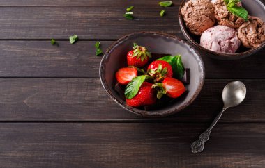 Bir kasede taze çilek, çikolata ve nane var. Yakınlarda, bir kasede farklı tatlarda üç kepçe dondurma bulunur. Tahta masadaki kasenin yanında bir kaşık duruyor..