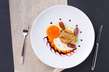 Bir gurme tabağı çeşitli soslar ve süslemelerle beyaz bir tabakta düzenlenir. Yemekte et, hurma ve turplar yer alıyor. Çatal ve bıçak karanlık bir masada, tabağın yanına konur..