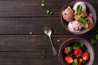 İki kase ahşap bir masada oturur. Birinin üzerinde nane yapraklı farklı dondurma çeşitleri var. Diğer kasede taze çilek var. Kâselerin yanında bir kaşık yatıyor..