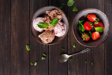 İki kase ahşap bir masada oturur. Bir kasede farklı tatlarda dondurma kepçeleri var. Diğer kase taze çileklerle dolu. Nane yaprakları üste konur.