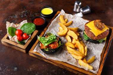 Peynirli bir hamburger, kızarmış patates ve taze salatanın yanında oturur. Kiraz domatesleri ve yapraklı yeşiller ahşap bir kasede düzenlenir. Soslar yakınlarda..