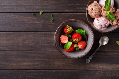 Tahta bir masanın üzerine iki kase konur. Bir kasede nane yapraklarıyla kaplı taze çilekler var. Diğer kasede farklı aromalı dondurmalar var, ayrıca naneyle süslenmiş..