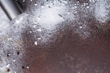 Bir tuzluk devrildi ve karanlık bir yüzeye tuz döktü. Sahne etrafa saçılmış tuz tanelerini gösteriyor. Biber taneleriyle karıştırılmış küçük tuz parçaları var..