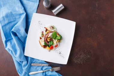 Çeşitli malzemelerle dolu bir yemek beyaz bir tabağa odaklıdır. Masa düzeninde mavi bir bez, bir çatal ve bir bıçak var. Çeşitli renkler ve yiyecek dokuları mevcuttur.