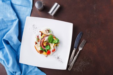 Beyaz bir tabağa yerleştirilmiş bir salata çeşitli sebzeler ve otlar içerir. Tabağın yanında gümüş çatal bıçak takımı ve arka planda bir tuzluk duruyor..