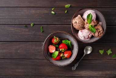 İki kase koyu ahşap bir masanın üzerinde. Bir kasede farklı tatlarda dondurma var. Diğer kasede taze çilek var. Dekorasyon için nane yaprakları üzerine yerleştirilir.