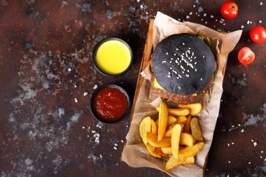 Bir siyah burger, ahşap bir tepside kızarmış patateslerin yanında oturur. İki küçük kase sos içeriyor ve yanına da vişneli domates konuyor. Ayar koyu bir masa yüzeyi..