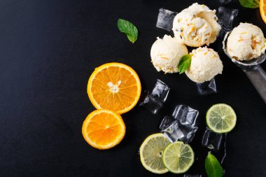 Portakal ve limon dilimlerinin bir karışımı dondurmalı kremalı turunçgil dondurmasının yanında duruyor. Sahne karanlık bir yüzeye kurulmuş, taze malzemeler ve tatlıyı vurguluyor..