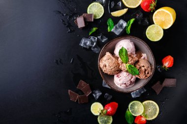 Bir kasede taze çilek, limon, nane ve çikolata parçalarıyla çevrili üç çeşit dondurma bulunur. Siyah yüzey, malzemelerin renkli düzenine zıtlık ekler.