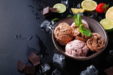 Dört kepçe dondurma, buz küpleri, limon ve limon dilimleri, taze çilekler ve çikolata parçalarıyla çevrili bir kasede. Sahne karanlık bir yüzeye kurulmuş..