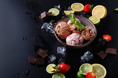 Bir kase çikolatalı ve çilekli dondurma içeriyor etrafında taze limon ve limon dilimleri, çikolata parçaları ve çilekler var. Buz küpleri sahneye serin bir dokunuş ekler..