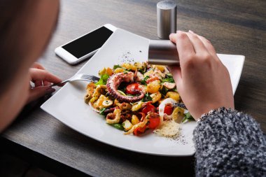 Bir kişi bir restoranda bir masada oturuyor, ahtapot, sebze ve makarnadan yapılmış bir salataya baharat ekliyor. Masanın yanında bir akıllı telefon var..
