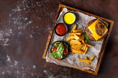 Bir tabakta peynir ve sebzeyle kaplı bir hamburger, yanında kızarmış patates ve sosla servis ediliyor. Yemekler parşömen kağıdında, tahta tepside..