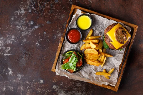 Bir tabakta peynir ve sebzeyle kaplı bir hamburger, yanında kızarmış patates ve sosla servis ediliyor. Yemekler parşömen kağıdında, tahta tepside..