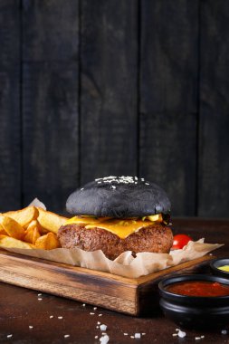 Peynirli bir siyah burger, altın kızartmalarla çevrili tahta bir tabakta kâğıt üzerinde oturur. Tabağın yanına küçük sos kaseleri konur. Arkaplanda koyu tahta paneller var..