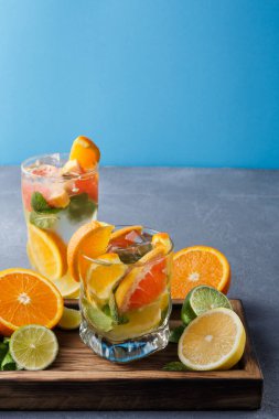 Turunçgil meyveleriyle dolu iki içecek ahşap bir tepsiye konur. Limon, limon ve portakal dilimleri bardakların etrafını sarar. Mavi arka plan meyve ve içeceklerin rengini artırır.