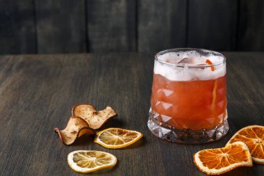 Renkli bir kokteyl ile dolu berrak bir bardak ahşap bir masada duruyor. İçkiye kurumuş limon ve portakal dilimleri eşlik ediyor. Buz küpleri bardakta görünür..