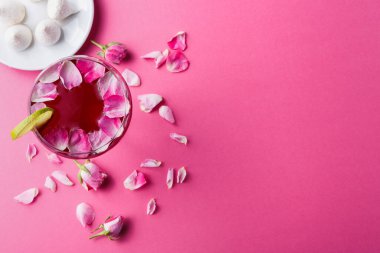 Bir içki parlak pembe bir yüzeye oturur. Bardak gül yapraklarıyla kaplı kırmızı bir içecekle dolu. Yakınlarda, bir tabakta küçük tatlılar var. Gül yaprakları içkinin etrafına saçılmış..