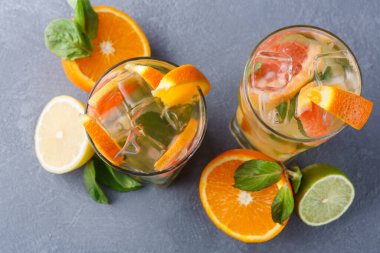 Turunçgil içecekleriyle dolu iki uzun bardak gri bir yüzeye oturur. İçeceklerde buz var ve portakal ve limon dilimleri ile süslenmiş. Bardakların etrafında taze nane yaprakları ve meyve parçaları var..