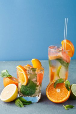 Buz, narenciye dilimleri ve taze nane yapraklarıyla dolu iki bardakta mavi bir masada oturuyorlar. Güneş ışığı meyve ve içeceğin renklerini ön plana çıkarıyor..