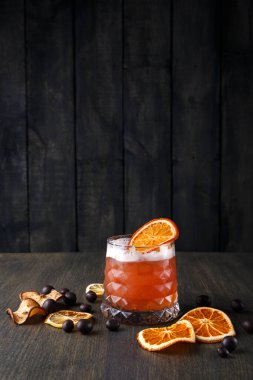 Bir kokteyl, etrafında portakal ve kurutulmuş meyve dilimleri olan ahşap bir masada oturur. İçkinin canlı bir rengi ve kenarında bir dilim turuncu var. Akşam ışığı sıcaklık katar..