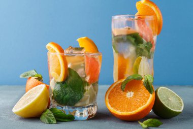 Renkli içeceklerle dolu iki bardak bir masada oturur. İçkilerde portakal ve limon dilimleri var. Nane yaprakları görünür. Bardakların etrafına biraz meyve konur..