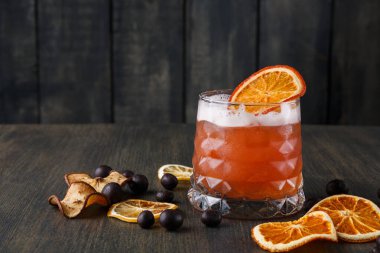 Buzlu, berrak bir bardakta kokteyl servis edilir. Kenarında bir portakal dilimi duruyor. Kurumuş meyve parçaları, tahta bir masanın üzerindeki camın etrafına saçılmış..