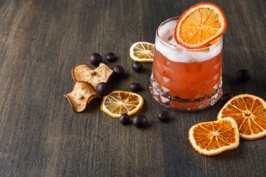 Sahne, üzerinde kurumuş portakal dilimi olan renkli bir içecekle dolu bir bardak gösteriyor. Camın etrafında kurumuş elma ve portakal dilimleri ve ahşap bir yüzey üzerinde koyu renkli meyveler var..