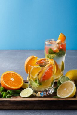 İki bardak narenciye içkisi ahşap tepside oturur. Portakal ve limon dilimleri nane yapraklarıyla düzenlenir. Arka plan mavi, parlak ve ferahlatıcı bir sahne yaratıyor..