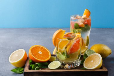 İki temiz bardakta taze meyve ve nane var. Portakal, limon ve kireç dilimleri ahşap bir tepside renkli bir görüntü oluşturuyor. Arka plan mavi..